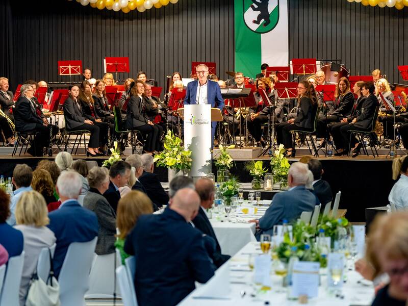 Eine Gruppe von Menschen sitzt bei einer Veranstaltung an Tischen und hört dem Bürgermeister bei seiner Rede zu, während im Hintergrund ein Blasorchester auf einer Bühne sitzt.