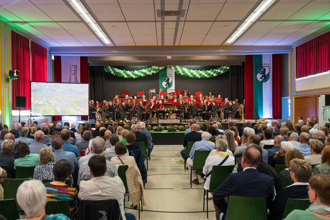 Ein Orchester spielt vor einem sitzenden Publikum in einem festlich dekorierten Saal mit Bannern und grünen Luftballons.