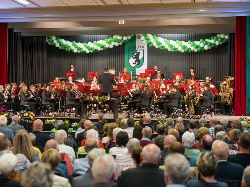 Auf dem Bild ist ein Blasmusikorchester in einer Halle zu sehen, das auf einer Bühne mit grün-weißer Dekoration vor einem sitzenden Publikum spielt.