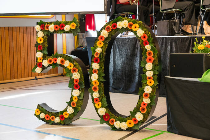 Das Bild zeigt große dekorative Ziffern "50", die mit grünem Moos und bunten Blumen geschmückt sind, aufgestellt in einem Innenraum mit Stühlen und einem Podium im Hintergrund.