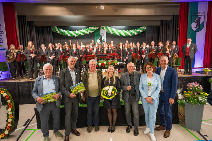 Auf dem Foto ist eine Gruppe von Menschen bei einer festlichen Veranstaltung zu sehen, wobei im Vordergrund einige Personen mit Blumen und Auszeichnungen stehen und dahinter eine größere Gruppe in formeller Kleidung mit Musikinstrumenten posiert, was auf ein Orchester oder eine musikalische Aufführung hindeutet.