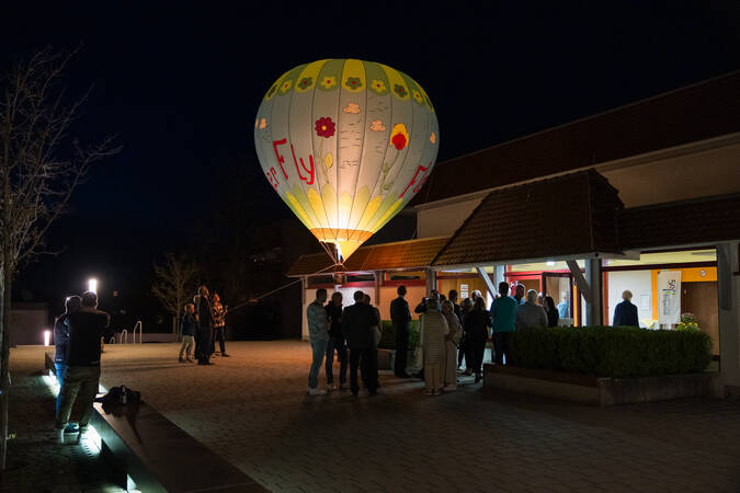 Ein beleuchteter Heißluftballon mit Blumendesign schwebt bei Nacht nahe einer Gruppe von Menschen, die sich vor einem Gebäude mit erleuchteten Fenstern versammelt haben.