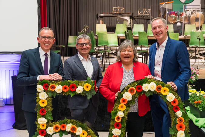 Vier Personen lächeln in die Kamera, während sie hinter einem großen, blumenverzierten Schriftzug stehen, vor dem Hintergrund einer Bühne mit grünen Stühlen.