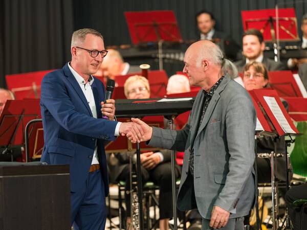 Zwei Männer schütteln sich auf einer Bühne vor einer Sitzkapelle die Hände, einer hält ein Mikrofon und beide lächeln.