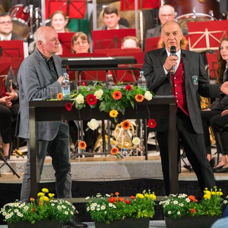 Zwei Männer stehen an einem Tisch mit Mikrofonen, umgeben von Blumenarrangements, während im Hintergrund eine Band sitzt.