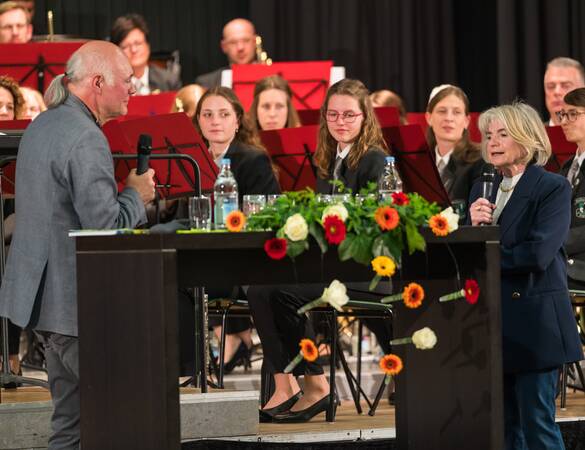 Zwei Personen sprechen an separaten Rednerpulten vor einem Orchester, das durch Blumenarrangements geschmückt ist.