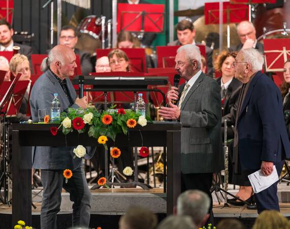 Drei Herren stehen auf einer Bühne bei einer Veranstaltung, zwei davon reden in Mikrofone und ein Orchester sitzt im Hintergrund.