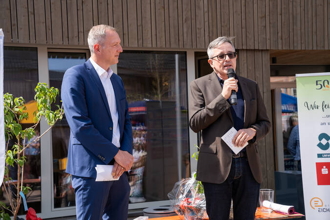 Zwei Männer, einer spricht in ein Mikrofon, während der andere zusehen scheint, stehen vor einem Publikum im Freien bei einer offensichtlichen Veranstaltung oder Feierlichkeit.