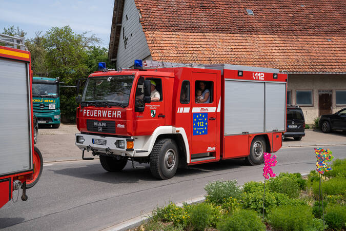 Ein rotes Feuerwehrfahrzeug fährt auf einer Straße neben einem Haus und einem parkenden Transporter, während ein Feuerwehrmann aus dem Fenster des Fahrzeugs schaut.