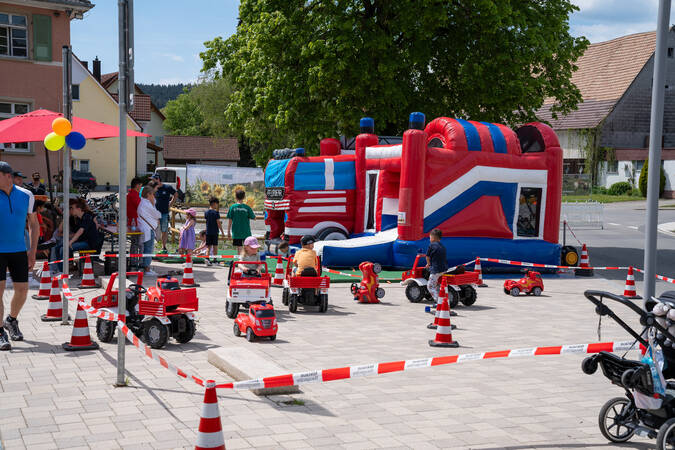 Ein belebter Außenbereich bei einem Fest mit Kindern, die auf einem abgesperrten Parcours mit Tretautos fahren, und einem großen aufblasbaren Spielgerät im Hintergrund.