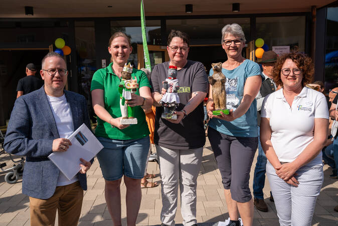 Fünf Personen stehen nebeneinander im Freien, lächeln und halten jeweils Trophäen in Form von Figuren.