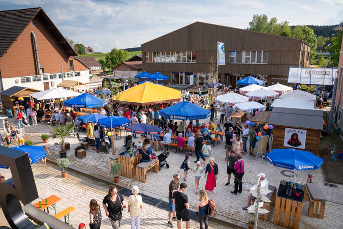 Es handelt sich um ein belebtes Straßenfest mit vielen Menschen, die zwischen verschiedenen Marktständen und unter Sonnenschirmen umhergehen und sich unterhalten, im Hintergrund ist ein modernes Gebäude sichtbar.