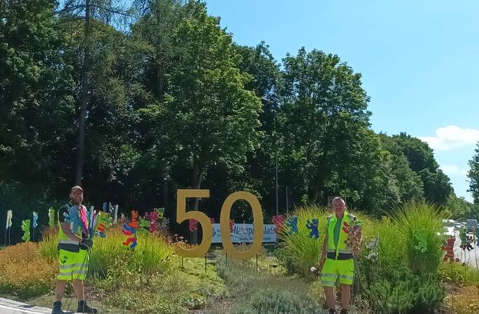 Das Bild zeigt zwei Personen in Warnwesten, die neben einer großen goldenen "50" stehen, umgeben von bunten Figuren und Pflanzen auf einer sonnigen Verkehrsinsel.