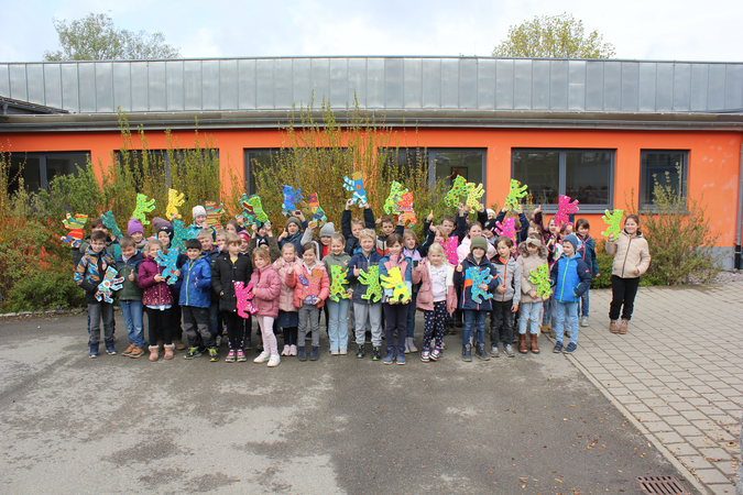 Eine Gruppe von Kindern steht vor einem Gebäude mit orangefarbener Fassade und hält bunte Holzbärenhoch.