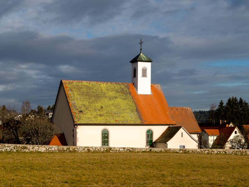 Im Vordergrund sieht man eine gemähte Wiese und im Hintergrund eine Kirche die von der Sonne angestrahlt wird. 