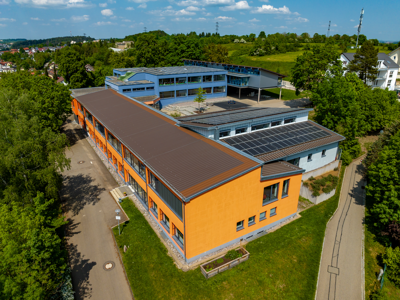 Luftaufnahme eines modernen Schulgebäudes mit orangefarbenen Wänden und Solarpanelen auf dem Dach, umgeben von Bäumen und einer Wiese.