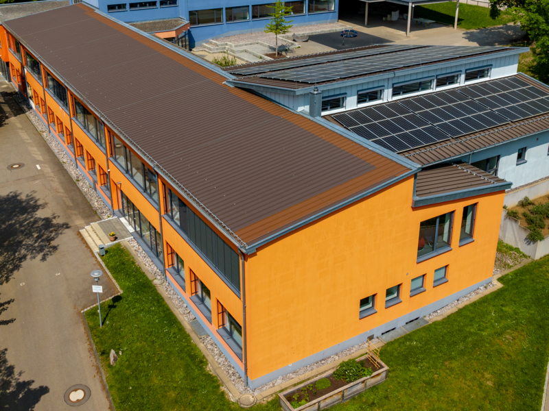 Luftaufnahme eines modernen Schulgebäudes mit orangefarbenen Wänden und Solarpanelen auf dem Dach, umgeben von Bäumen und einer Wiese.