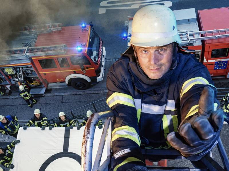 Ein Feuerwehrmann steht auf einer Leiter und reicht eine Hand, andere Feuerwehrmänner stehen mit einem Sprungtuch im Hintergrund 