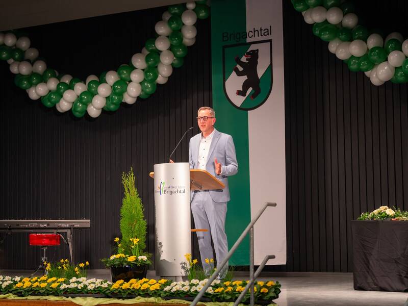 Der Bürgermeister steht an einem Rednerpult und spricht, während im Hintergrund Dekorationen in Grün-Weiß und ein Emblem mit einem Bären zu sehen sind.