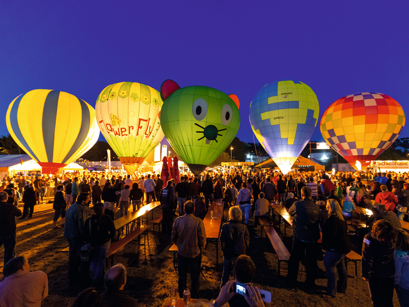 Ein abendliches Ballonfestival mit leuchtenden Heißluftballonen unterschiedlicher Farben und Designs, umgeben von einer Menschenmenge.