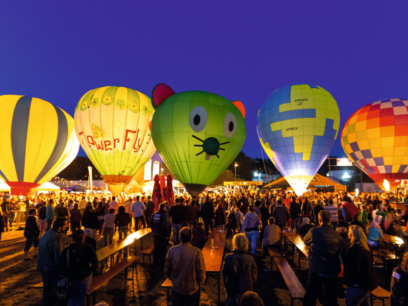 Ein abendliches Ballonfestival mit leuchtenden Heißluftballonen unterschiedlicher Farben und Designs, umgeben von einer Menschenmenge.