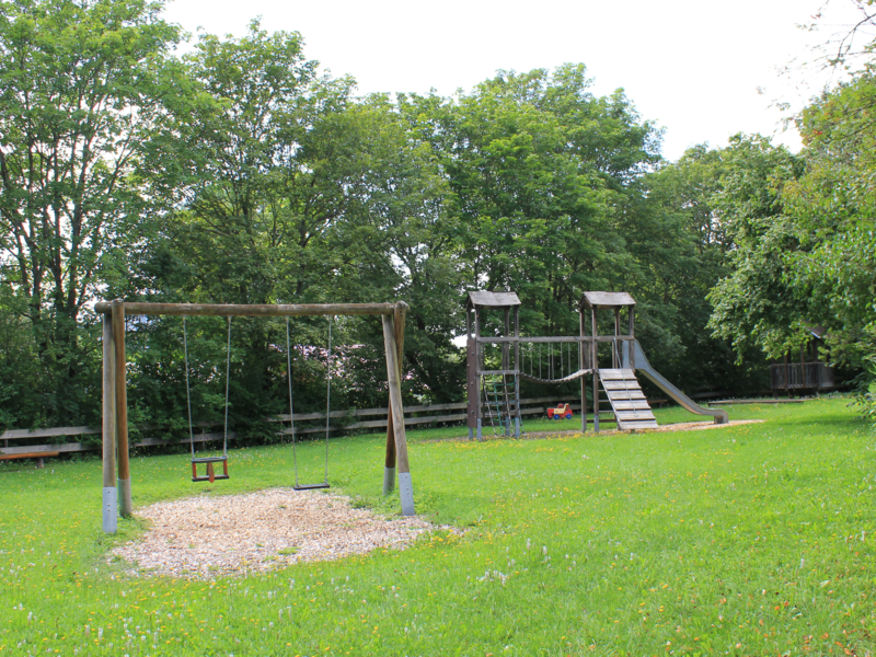 Ein Kinderspielplatz mit einer Schaukel und einer Rutsche, umgeben von grünen Bäumen und einer Wiese.