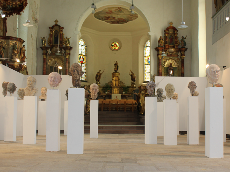 In einem Kirchenraum sind mehrere Büsten auf weißen Sockeln ausgestellt, im Hintergrund sind Kirchenbänke und farbige Glasfenster zu sehen.