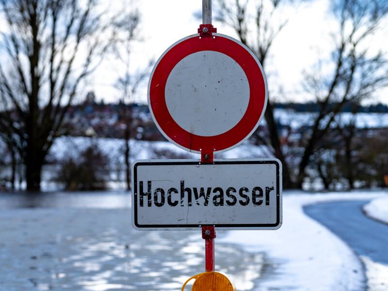 Ein Verkehrsschild mit der Aufschrift "Hochwasser" steht vor einer überfluteten Straße mit Eisflächen und Schnee sowie Bäumen im Hintergrund.