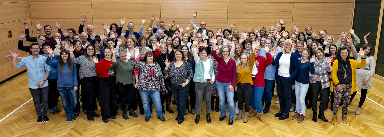 Die Mitarbeiter der Gemeinde winken in die Kamera und lachen, im Hintergrund ist das innere des Dorfhauses zu sehen. 