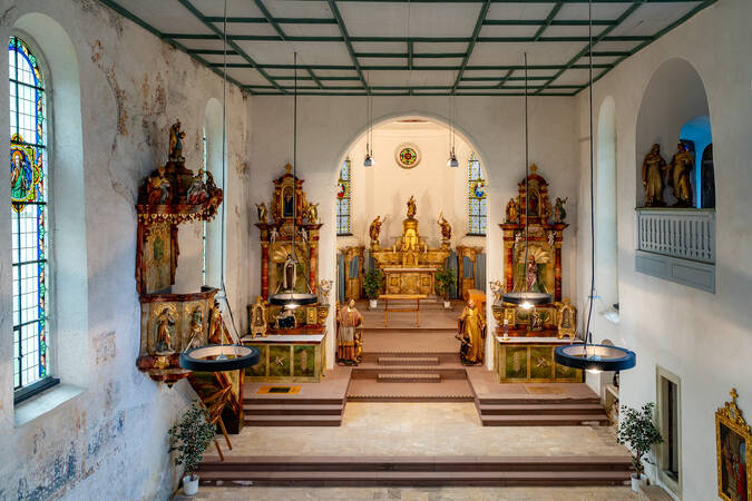 Das Bild zeigt den Innenraum einer Kirche mit einem zentralen Altar, Seitenaltären, bunten Kirchenfenstern und religiösen Figuren.