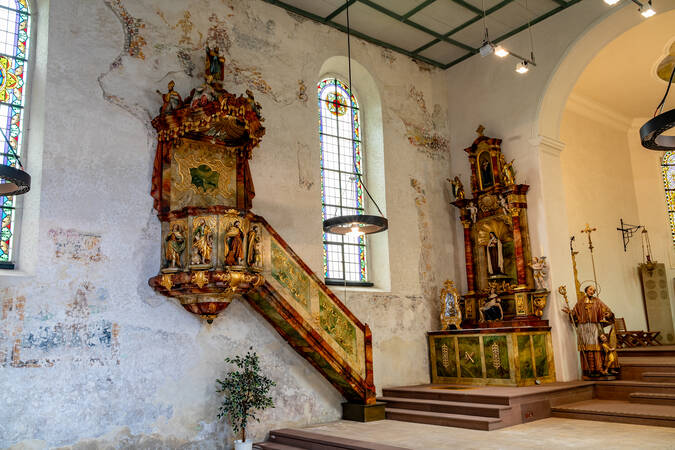 Das Bild zeigt den Innenraum einer Kirche mit einem zentralen Altar, Seitenaltären, bunten Kirchenfenstern und religiösen Figuren.