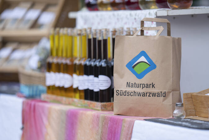 Eine Papiertüte mit dem Aufdruck "Naturpark Südschwarzwald" steht vor unscharfem Hintergrund mit Flaschen und anderen Marktprodukten auf einer Auslage.