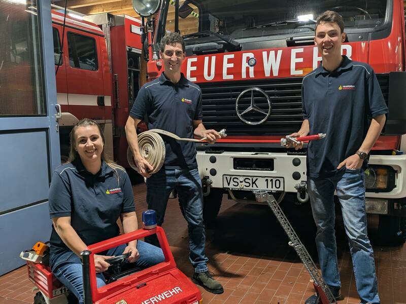 Drei Personen in Feuerwehruniformen stehen vor einem großen Feuerwehrwagen, wobei eine Person auf einem kleinen Spielzeug-Feuerwehrauto sitzt und die anderen zwei Feuerwehrschläuche halten.