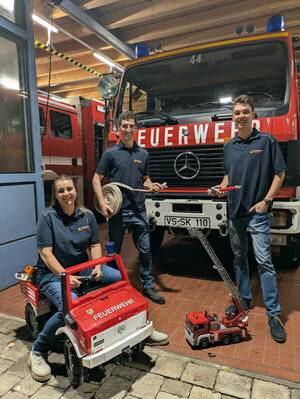 Drei Personen in Feuerwehruniformen stehen vor einem großen Feuerwehrwagen, wobei eine Person auf einem kleinen Spielzeug-Feuerwehrauto sitzt und die anderen zwei Feuerwehrschläuche halten.