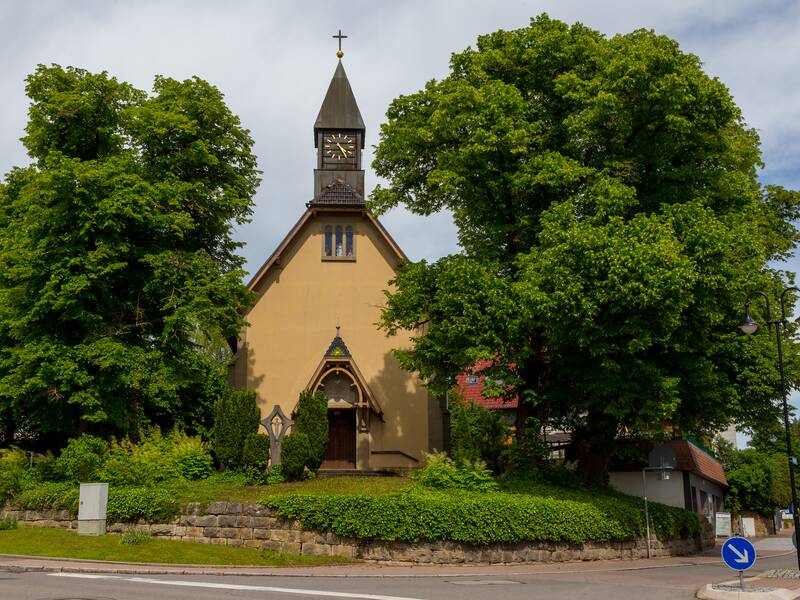 Eine kleine Kirche mit spitzen Turmdach und Uhr, eingebettet zwischen üppigen grünen Bäumen, steht neben einer Straße mit einer Straßenlaterne und Verkehrsschildern im Vordergrund.