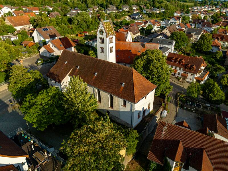 Luftaufnahme einer Kirche mit weißer Fassade und rotem Dach, eingebettet in eine ländliche Ortschaft mit grünen Bäumen und traditionellen Häusern bei Tageslicht.