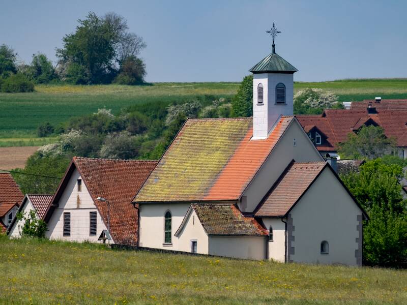 Eine ländliche Szenerie mit einer kleinen Kirche und umliegenden Wohnhäusern, eingebettet in eine Landschaft mit grünen Feldern und Bäumen im Hintergrund.