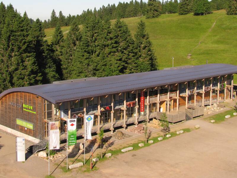 Ein langgezogenes Gebäude mit Holzfassade und einem gewölbten Dach steht im Vordergrund, umgeben von grünen Bäumen und einer weitläufigen Landschaft mit Grashängen unter blauem Himmel.