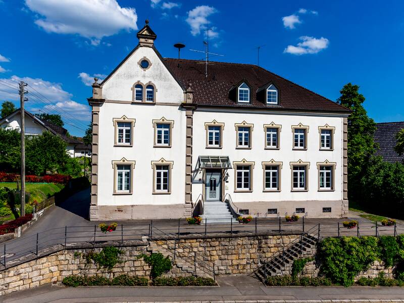 Das Bild zeigt ein zweistöckiges, weißes Gebäude mit einem Giebeldach und Zierleisten um die Fenster, gelegen an einer Straße mit einer Steinmauer und gepflegten Beeten.