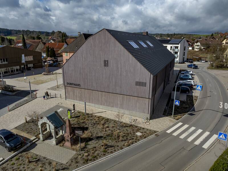 Ein modernes, mit Holz verkleidetes Gebäude mit der Aufschrift "DORFHAUS" steht an einer Straßenecke in einer ländlichen Siedlung bei Tageslicht.