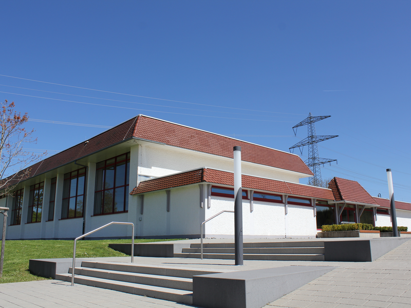 Ein modernes, eingeschossiges Gebäude mit rotem Dach und großen Fenstern unter blauem Himmel, vor dem sich eine gepflasterte Fläche mit Treppen und Geländern befindet.