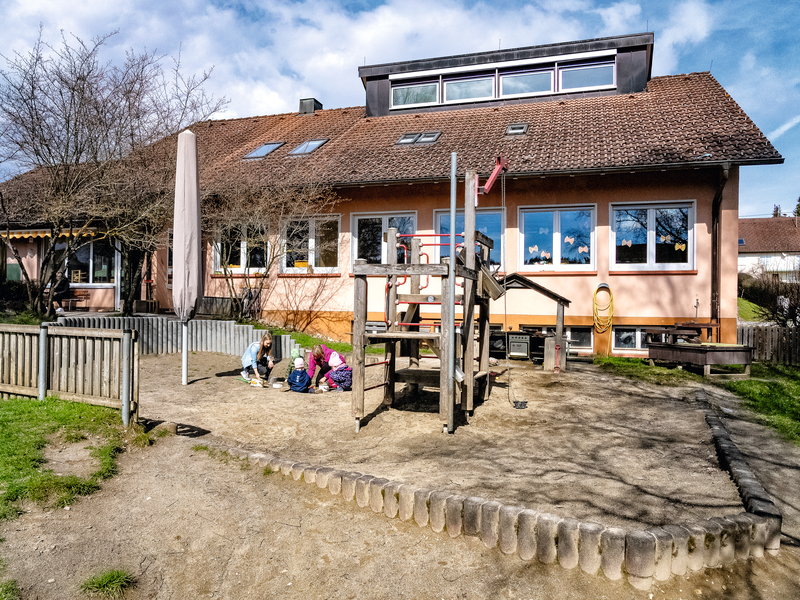 Ein Sandkasten mit Spielgerät im Vordergrund, im Hindergrund die Kita am Vorberg