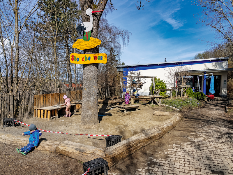 Im Hintergrund die Kita Storchennest unter blauem Himmel. Im Vordergrund das Eingangsschild mit Storch.