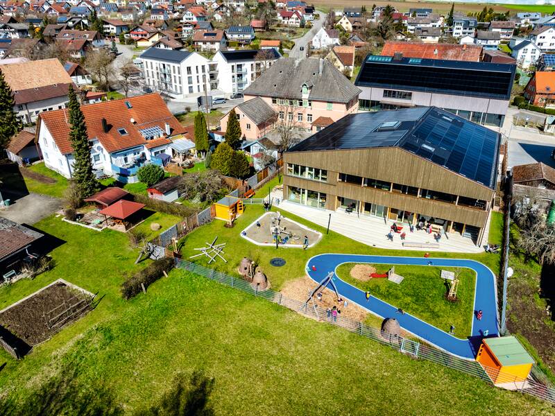 Luftaufnahme einer städtischen Szenerie mit einem modernen Gebäude und angrenzendem Spielplatz neben traditionellen Wohnhäusern.