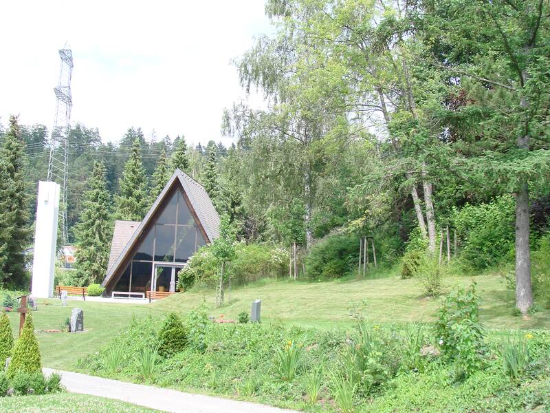 Ein modernes, spitz zulaufendes Gebäude mit großen Fenstern steht umgeben von grünen Bäumen und einer gepflegten Rasenfläche, neben einem weißen Turm und einer metallischen Maststruktur.