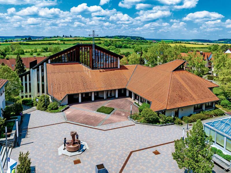 Ein großes Gebäude mit terracottafarbenem Ziegeldach und einer Glasfront, umgeben von Bäumen und Wiesen, bei sonnigem Wetter.