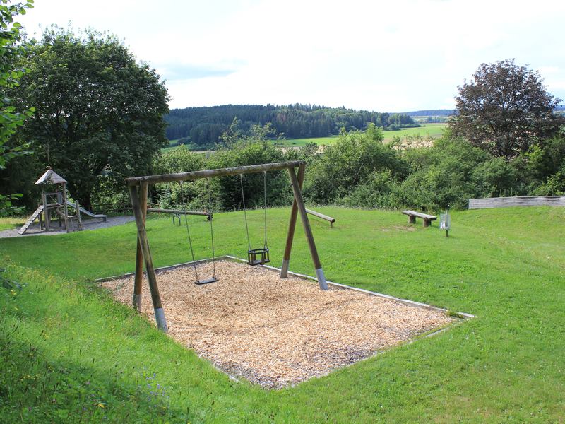 Ein Spielplatz mit Schaukel, Rutsche und Wippe auf grüner Wiese mit blauem Himmel und Wald im Hintergrund.