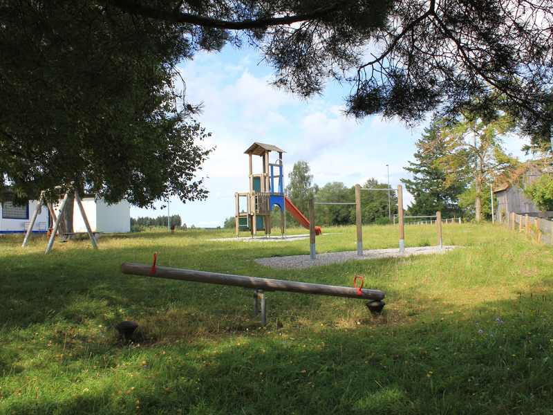 Eine Wippe, eine Reckstange, ein Spielturm mit Rutsche und eine Schaukel auf grüner Wiese unter blauem Himmel.