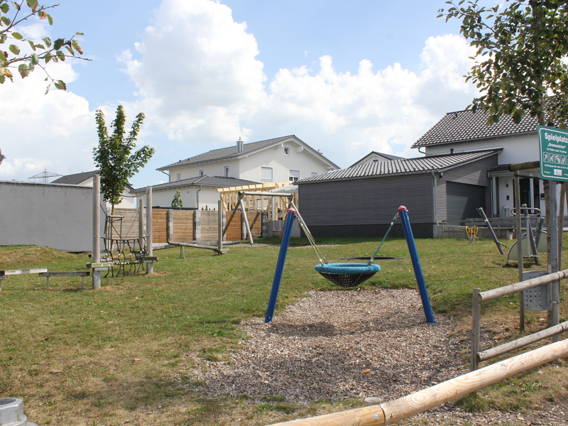 Ein kinderfreundlich gestalteter Spielplatz mit Schaukeln, Rutschen und anderen Spielgeräten, umgeben von einem Zaun und Wohnhäusern im Hintergrund.