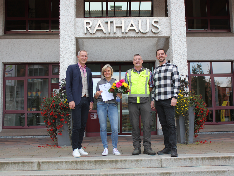 Vor dem Rathausgebäude stehen vier lächelnde Personen, eine Frau hält Blumen und eine Urkunde in der Hand.